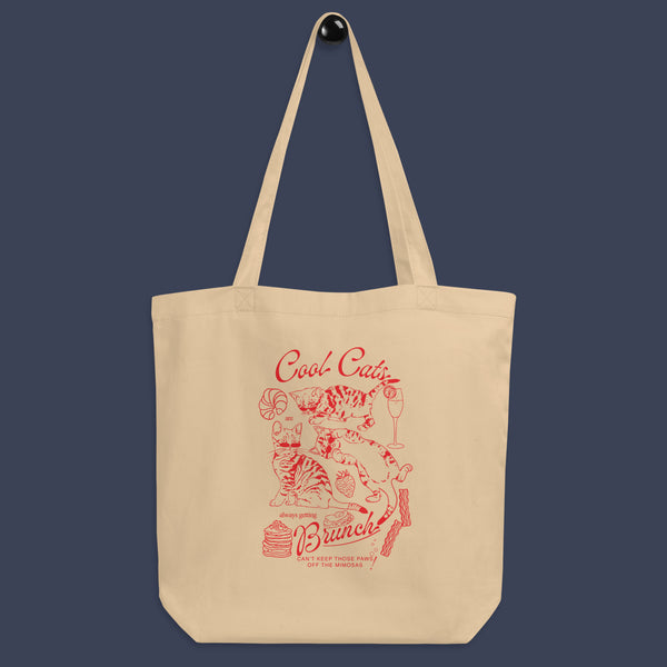 a picture of a tote bag hung on a simple black peg. The tote bag is a natural oyster colour with a red line art design of kittens and brunch food with the phrase cool cats are always getting brunch on it.