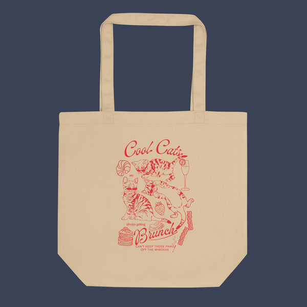 a picture of a tote bag  laid flat against a dark blue background. The tote bag is a natural oyster colour with a red line art design of kittens and brunch food with the phrase cool cats are always getting brunch on it.