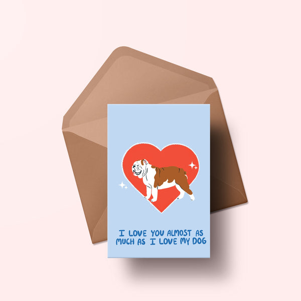 image of a greetings card featuring an illustration of an english bulldog with a red heart behind it. The background of the card is a light blue and beneath the heart and dog are the words I love you almost as much as my dog in handwritten lettering in a darker blue shade. Behind the card is an envelope made of a recycled kraft material.