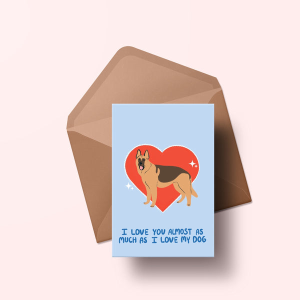 image of a greetings card featuring an illustration of a german shepherd with a red heart behind it. The background of the card is a light blue and beneath the heart and dog are the words I love you almost as much as my dog in handwritten lettering in a darker blue shade. Behind the card is an envelope made of a recycled kraft material.