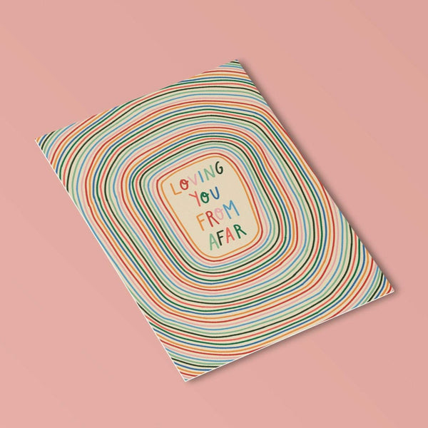 image of a postcard with the words loving you from afar handwritten in alternating rainbow text. the background consists of the same repeating rainbow colours in concentric circles. the background is a cream colour.