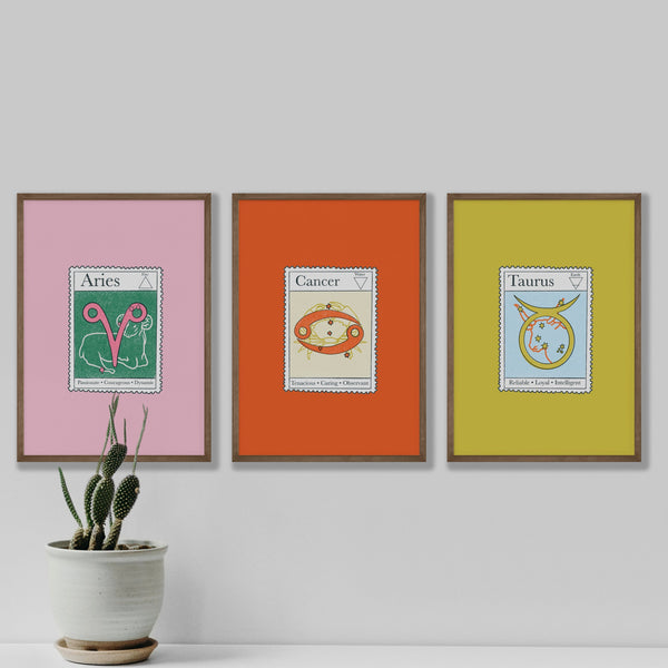 Image of three framed prints each with a star sign inspired postage stamp illustration. The frames are on a white wall. There is a small cactus plant in a white ceramic pot in the foreground.