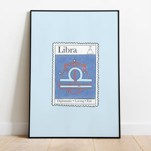 Image of a framed art print leaning against the wall. The art print itself is a postage stamp representing the star sign of Libra. The print is bright and colourful in shades of blues and red.