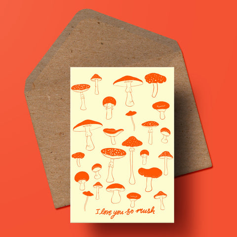 image of a greetings card featuring a selection of illustrated toadstools with the handwritten text 'i love you mush' underneath. beneath the card is a recycled kraft envelope.