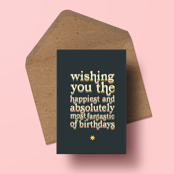 image of a birthday card in 70s rainbow typography that reads wishing you the happiest and absolutely most fantastic of birthdays. the card is resting on top of a recycled kraft envelope.