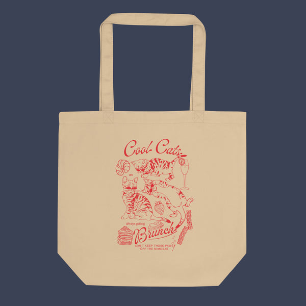 a picture of a tote bag  laid flat against a dark blue background. The tote bag is a natural oyster colour with a red line art design of kittens and brunch food with the phrase cool cats are always getting brunch on it. 