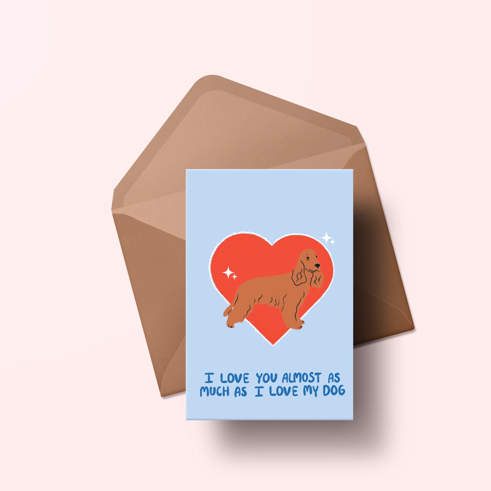 image of a greetings card featuring an illustration of a cocker spaniel with a red heart behind it. The background of the card is a light blue and beneath the heart and dog are the words I love you almost as much as my dog in handwritten lettering in a darker blue shade. Behind the card is an envelope made of a recycled kraft material.