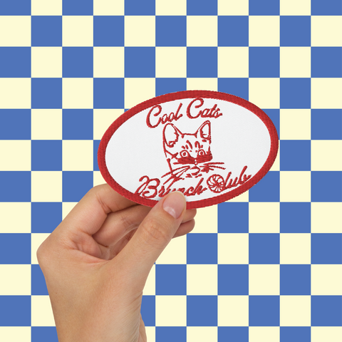 image of a hand holding an embroidered patch with the words cool cats brunch club on it in bright red. In the middle of the patch is the head of a kitten wearing sunglasses. the C of the 'club' is made out of a croissant
