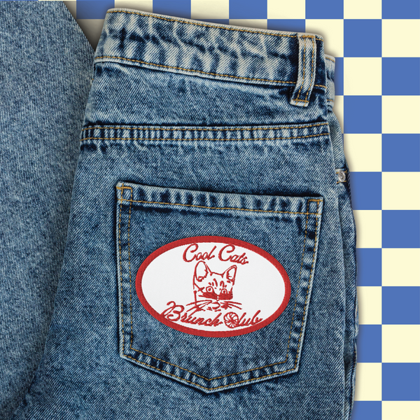 image of an embroidered patch sewn onto the back pocket of a pair of jeans with the words cool cats brunch club on it in bright red. In the middle of the patch is the head of a kitten wearing sunglasses. the C of the 'club' is made out of a croissant