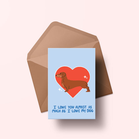 image of a greetings card featuring an illustration of a dachshund with a red heart behind it. The background of the card is a light blue and beneath the heart and dog are the words I love you almost as much as my dog in handwritten lettering in a darker blue shade. Behind the card is an envelope made of a recycled kraft material.
