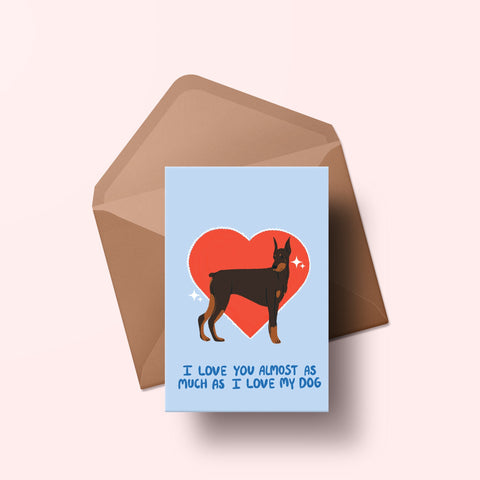 image of a greetings card featuring an illustration of a doberman with a red heart behind it. The background of the card is a light blue and beneath the heart and dog are the words I love you almost as much as my dog in handwritten lettering in a darker blue shade. Behind the card is an envelope made of a recycled kraft material.