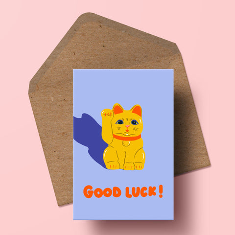 image of a greetings card featuring a golden lucky cat a 'maneki neko' with the words 'good luck!' underneath in vibrant red handwritten text. the background of the card is a pale cornflower blue. behind the card is a recycled kraft envelope.