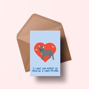 image of a greetings card featuring an illustration of a frenchie with a red heart behind it. The background of the card is a light blue and beneath the heart and dog are the words I love you almost as much as my dog in handwritten lettering in a darker blue shade. Behind the card is an envelope made of a recycled kraft material.