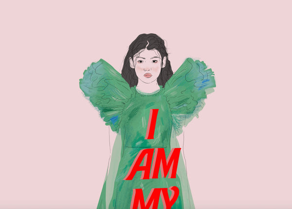 a zoomed in look at an illustration print of a young woman wearing a full length green dress with the words i am my own muse on it. the background is a light dusky pink.