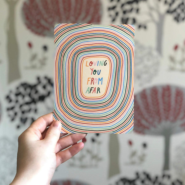 image of a hand holding a  postcard with the words loving you from afar handwritten in alternating rainbow text. the background consists of the same repeating rainbow colours in concentric circles. the background is a cream colour