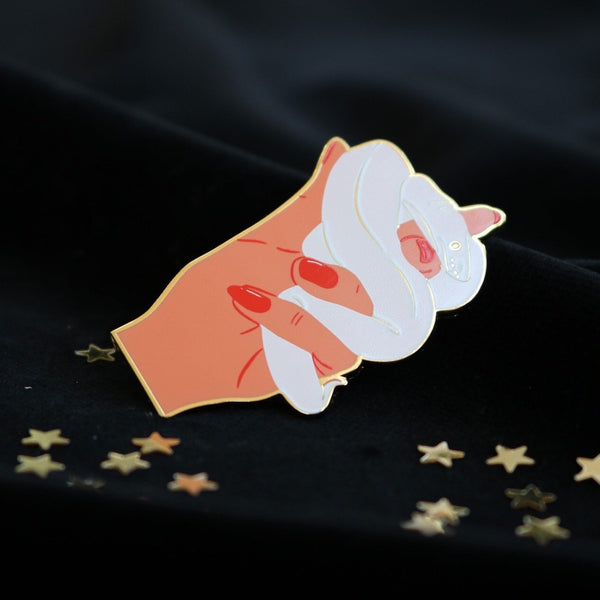 image of an enamel pin of a hand holding a white snake. the pin is on a black velvet background which is dotted with confetti stars. the light is shining off the pin