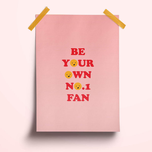 A typography print featuring the words 'be your own no.1 fan' the os are replaced with yellow smiley faces. The text is in a 70s style font in red. the background is a mid pink