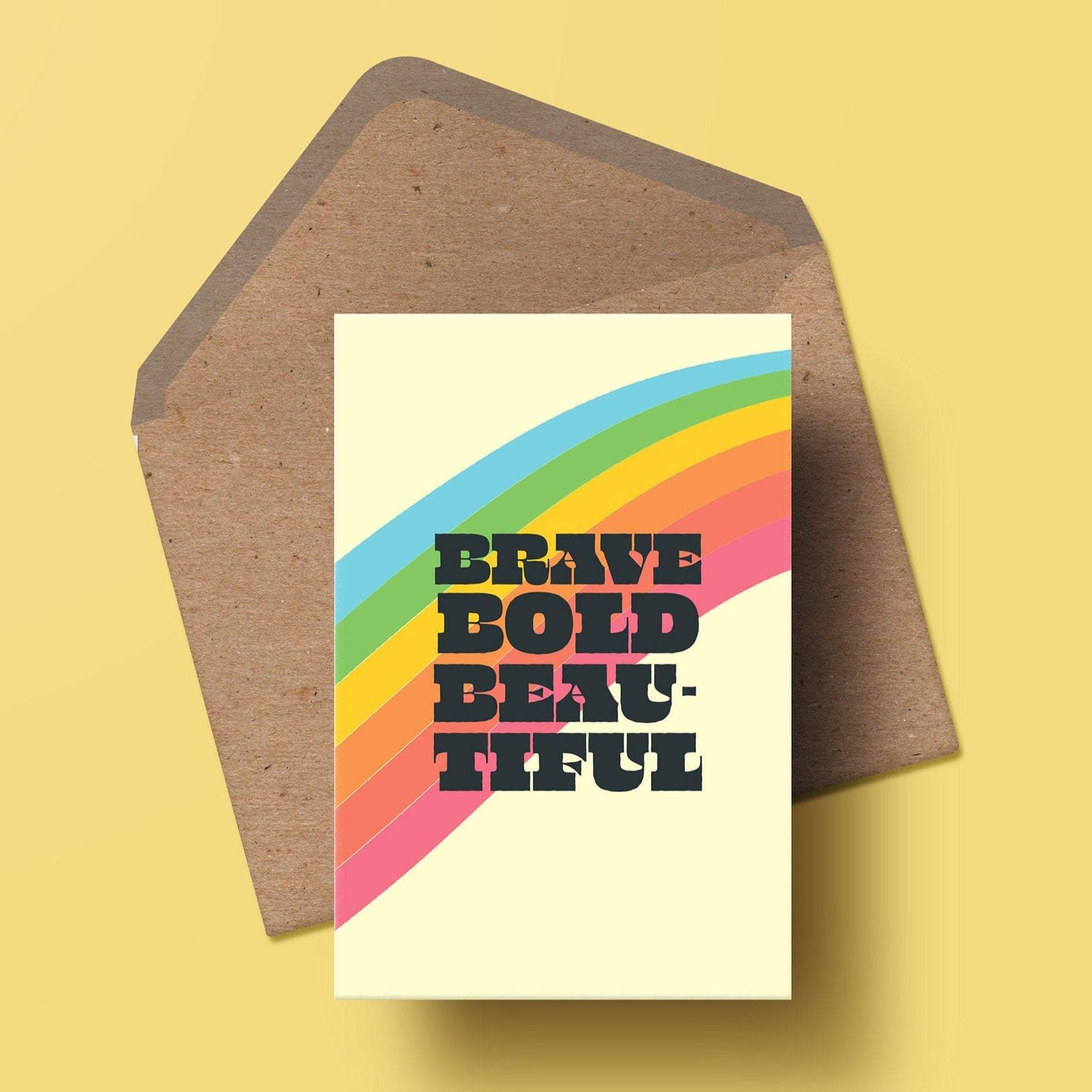 an a6 greetings card with the words brave, bold, beautiful in a bold black text, behind them is a rainbow stripe. there is a matching kraft envelope behind the card.