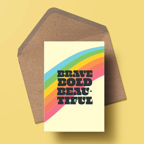 an a6 greetings card with the words brave, bold, beautiful in a bold black text, behind them is a rainbow stripe. there is a matching kraft envelope behind the card.