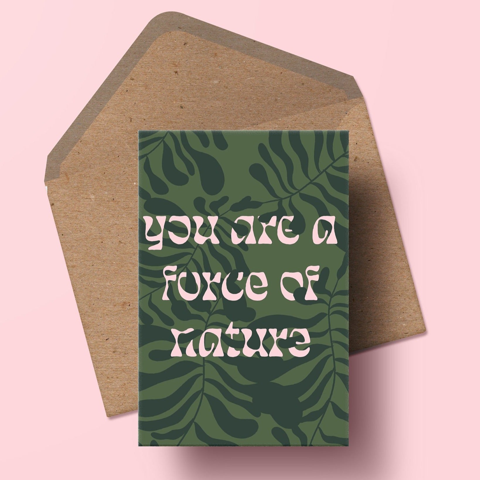 image of a greetings card featuring the words you are a force of nature in pale pink. the background is a medium green with a pattern of dark green overlapping leaves. the card is sat on top of a kraft envelope.