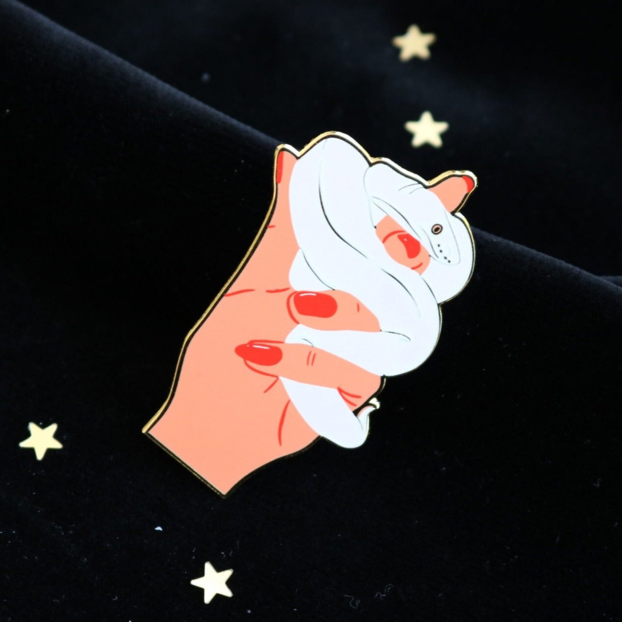 image of an enamel pin of a hand holding a white snake. the pin is on a black velvet background which is dotted with confetti stars
