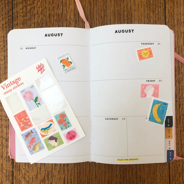 a sticker sheet featuring a selection of vintage inspired postal stamps. some of the stickers have been used in a planner which is open on a wooden table.
