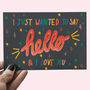 image of a hand holding a postcard with the handwritten words 'I just wanted to say hello and I love you' with rainbow accents on a dark navy background.