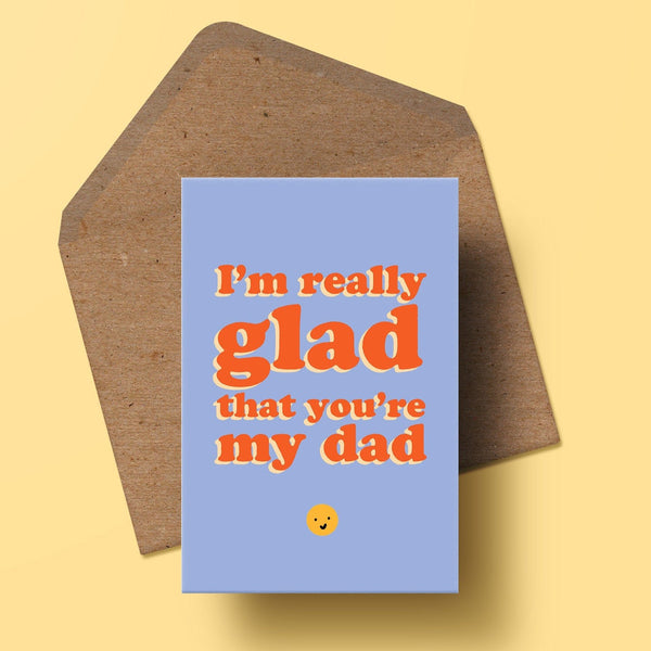 Image of a greetings card featuring the words I'm really glad that you're my dad in red type with a cream drop shadow. the background of the card is a medium blue colour and there is a small smiley face underneath the text. the card is presented with a recycled kraft envelope behind it.