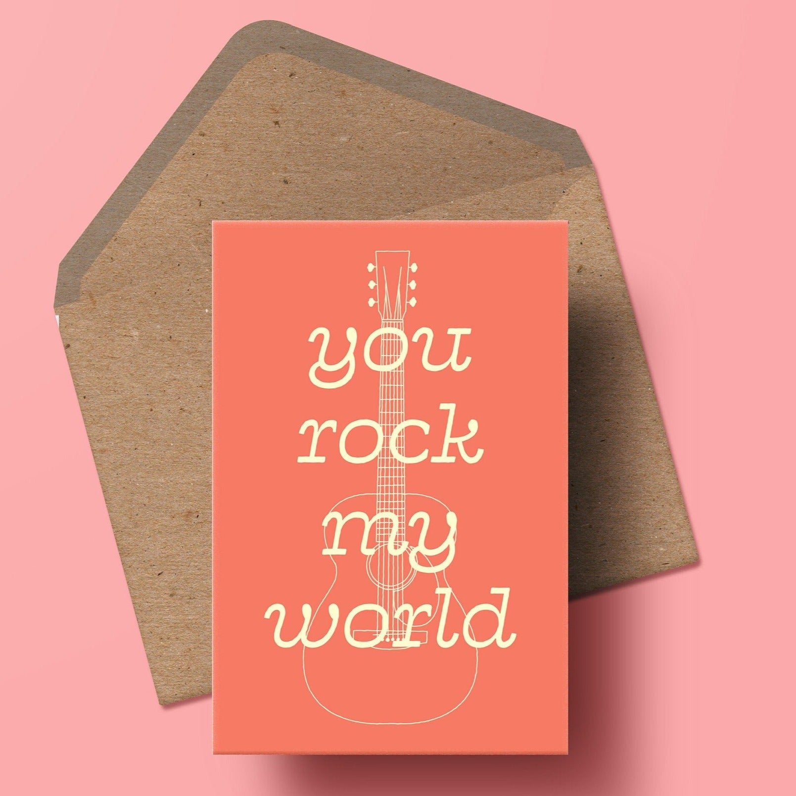 image of a greetings card featuring the words you rock my world in cream overlaid onto an illustration of an acoustic guitar also in cream. the background is a muted coral colour. beneath the card is a kraft envelope.