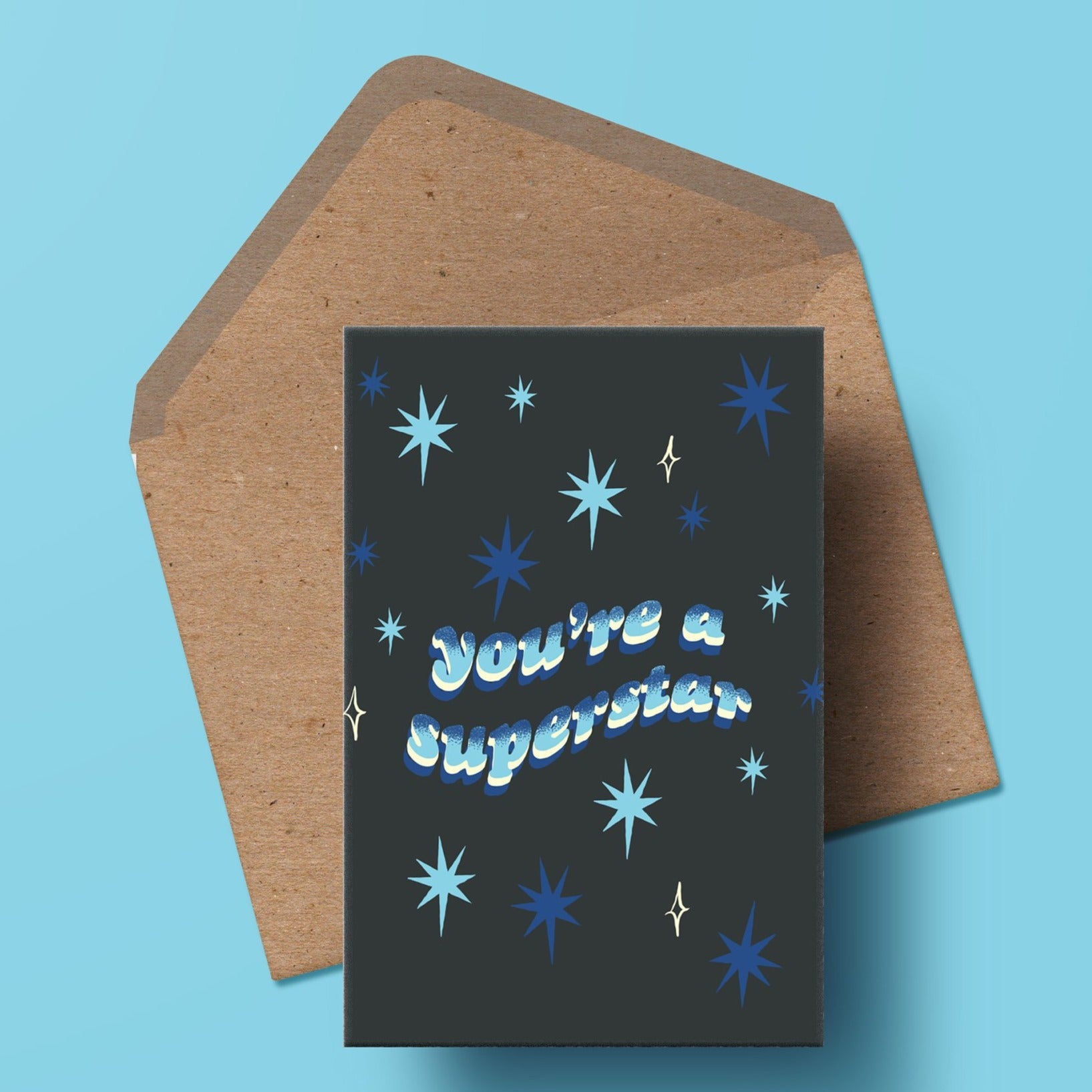 image of a greetings card featuring the words you're a superstar in a wavy 70s font with a hand drawn blue stipple gradient. the background is a deep navy with a starry motif. the card is sat on top of a kraft envelope.