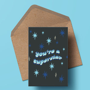 image of a greetings card featuring the words you're a superstar in a wavy 70s font with a hand drawn blue stipple gradient. the background is a deep navy with a starry motif. the card is sat on top of a kraft envelope.