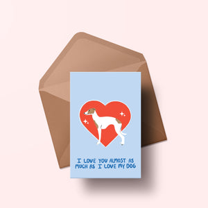 image of a greetings card featuring an illustration of an italian greyhound with a red heart behind it. The background of the card is a light blue and beneath the heart and dog are the words I love you almost as much as my dog in handwritten lettering in a darker blue shade. Behind the card is an envelope made of a recycled kraft material.
