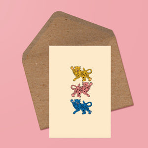 image of a greetings card featuring three leopards facing alternate directions with fur colours of yellow, pink and blue respectively. behind the card is a recycled kraft envelope