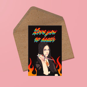 image of a card featuring an illustration of elaine from the love witch holding a bloody knife. behind her are stylised flames. the words love you to death in handwritten, rainbow cursive are above her. the card is presented with a recycled kraft envelope behind