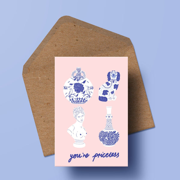 image of a greetings card featuring illustrations of two antique vases, a ceramic staffordshire dog and a marble bust. the background of the card is a pale pink. the words 'you're priceless' are written beneath. underneath the card is a recycled kraft envelope.