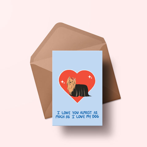 image of a greetings card featuring an illustration of a yorkie with a red heart behind it. The background of the card is a light blue and beneath the heart and dog are the words I love you almost as much as my dog in handwritten lettering in a darker blue shade. Behind the card is an envelope made of a recycled kraft material.